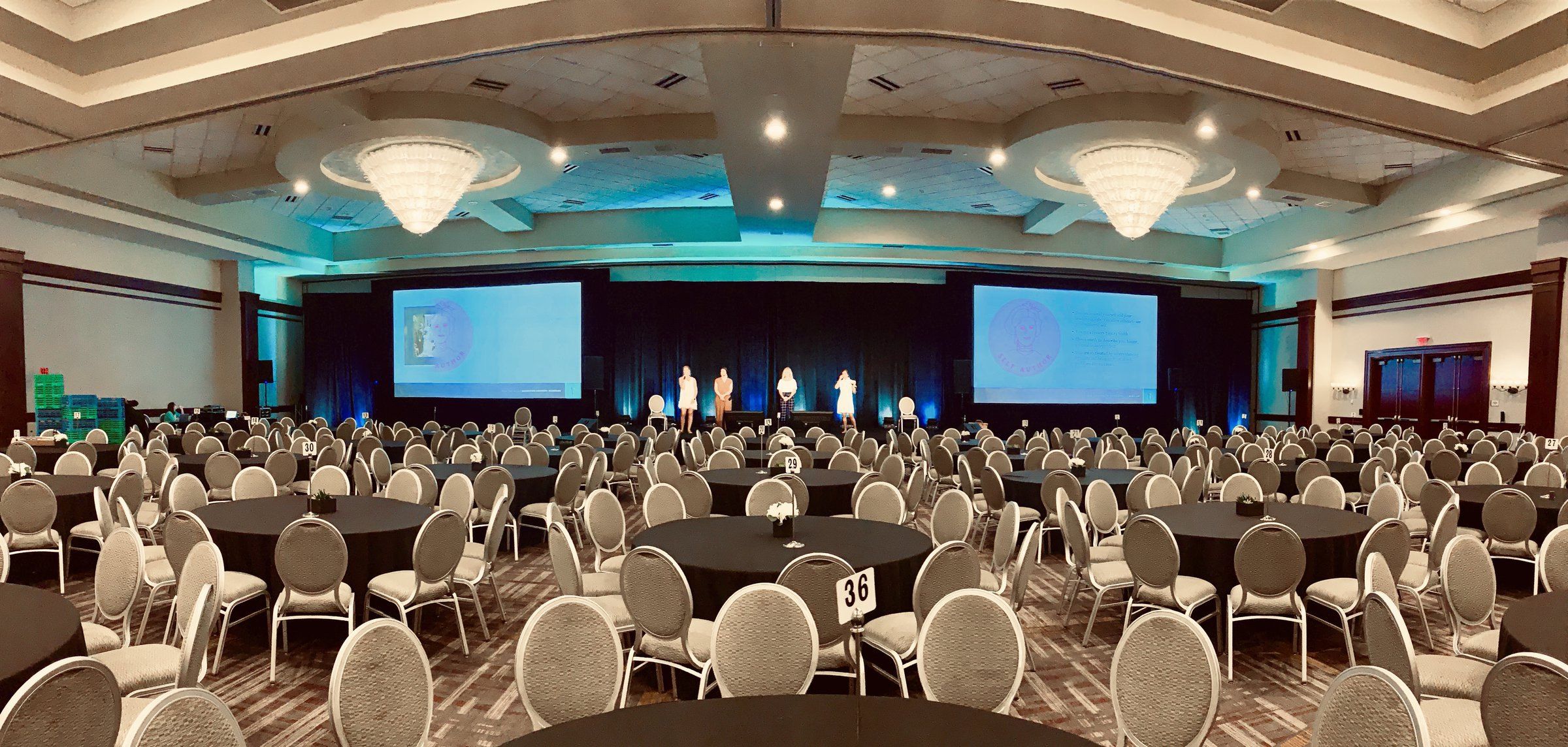 A large ballroom laid out with round tables facing a stage and twin screens